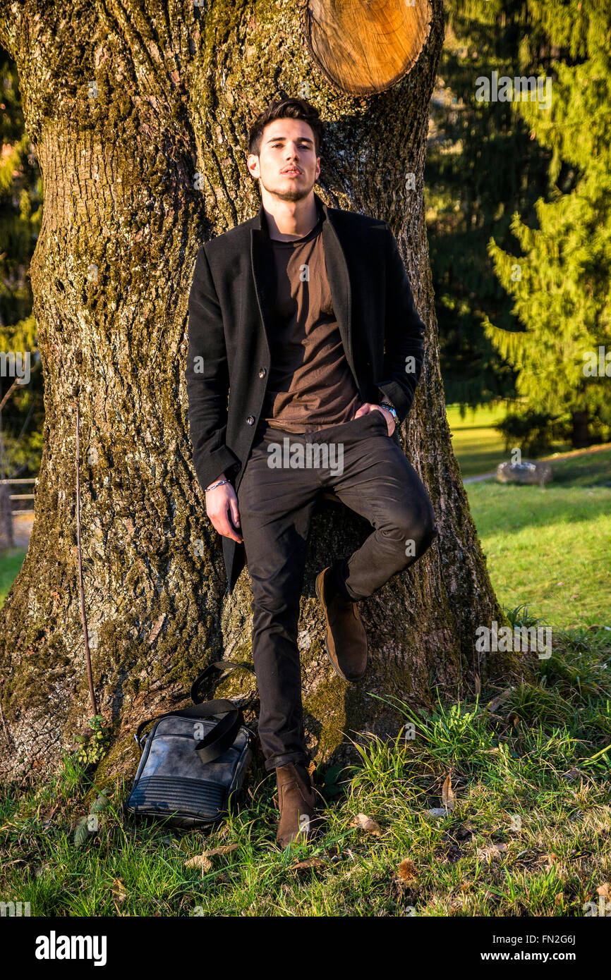 Bel giovane uomo appoggiato contro l'albero, guardando la telecamera, in una giornata di sole che indossa un mantello nero Foto Stock