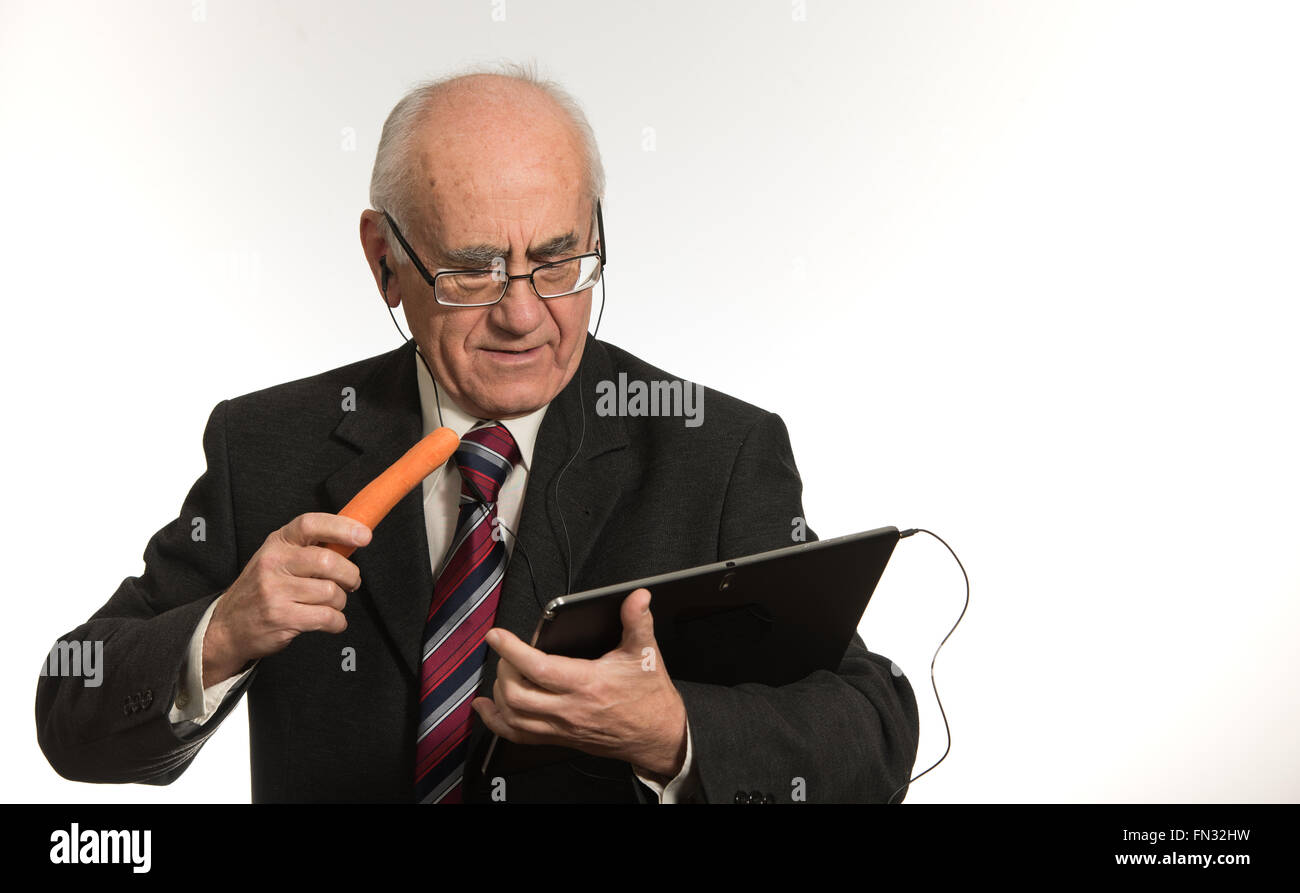 Vecchio imprenditore senior lavora con nero computer tablet, indossando il business suit, mangiando la carota Foto Stock Vecchio imprenditore senior lavora con nero computer tablet, indossando il business suit, mangiando la carota Foto Stock