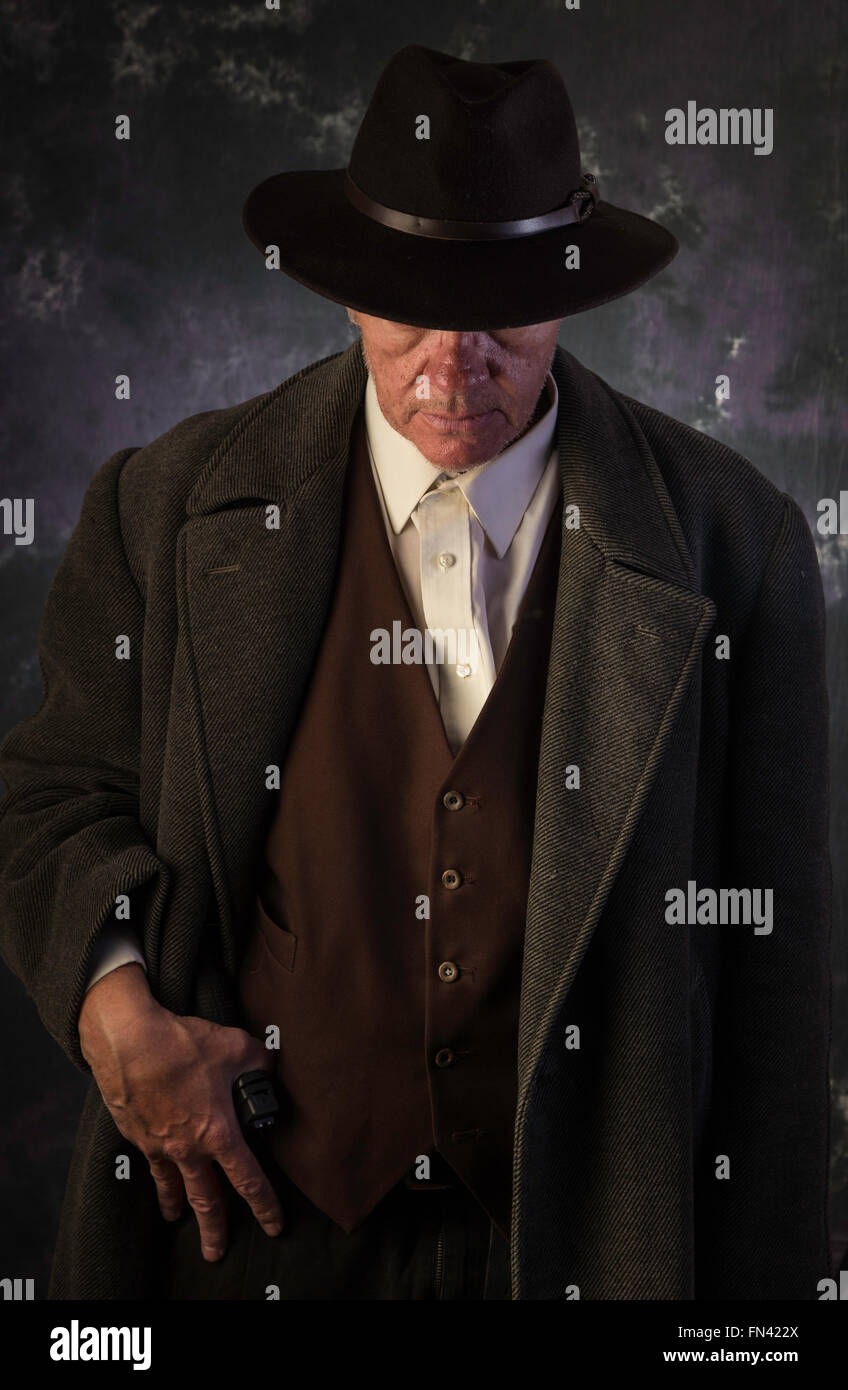 Sinister cerca uomo che indossa soprabito e il cappello con una mano sul belted pistola contro sfondo verticale in condizioni di scarsa illuminazione tasto Foto Stock