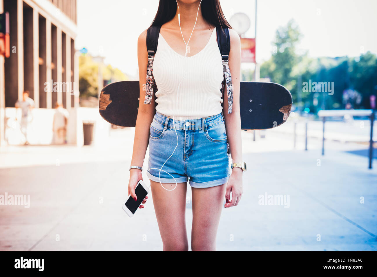 Dal collo in giù in vista della giovane donna con lo skateboard e zainetto, tenendo in mano un telefono intelligente e ascolto di musica con cuffie - Foto Stock