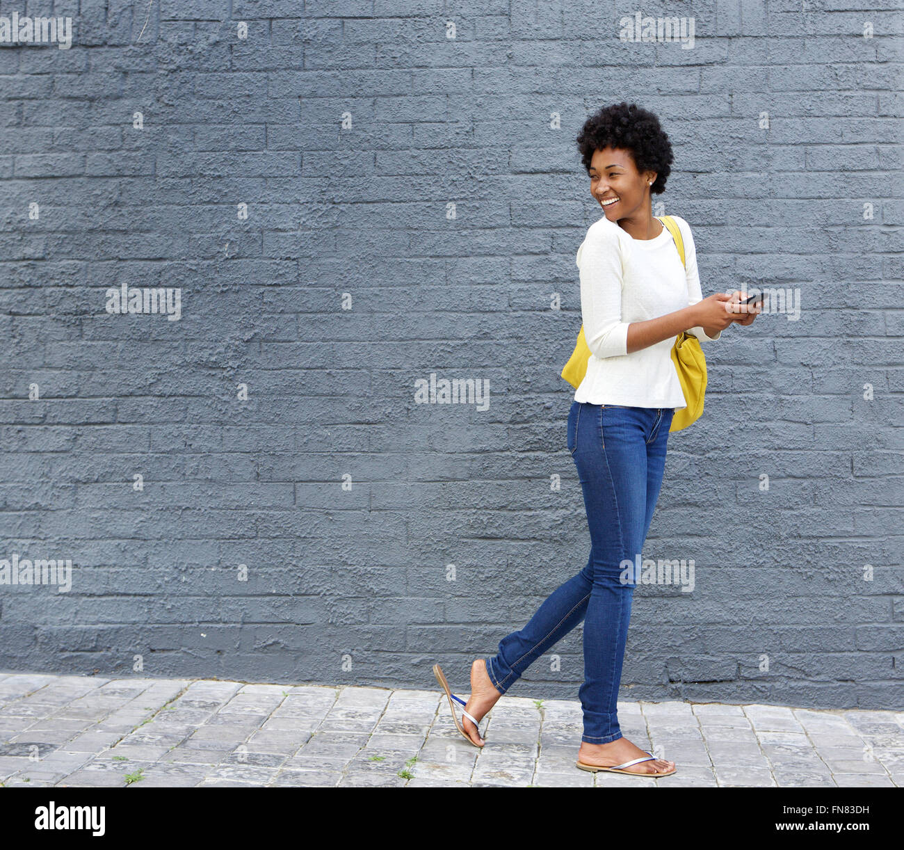 A piena lunghezza Ritratto di un felice giovane donna africana con un telefono cellulare a piedi sulla strada e guardando sopra la sua spalla Foto Stock