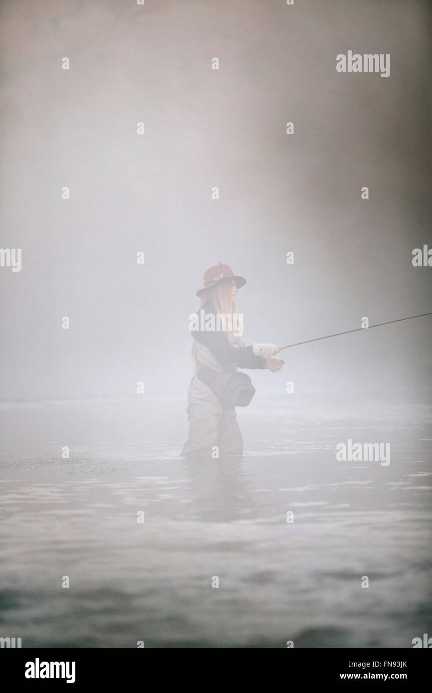 Una donna pescatore pesca a mosca, in piedi di waders a coscia in acque profonde. Foto Stock