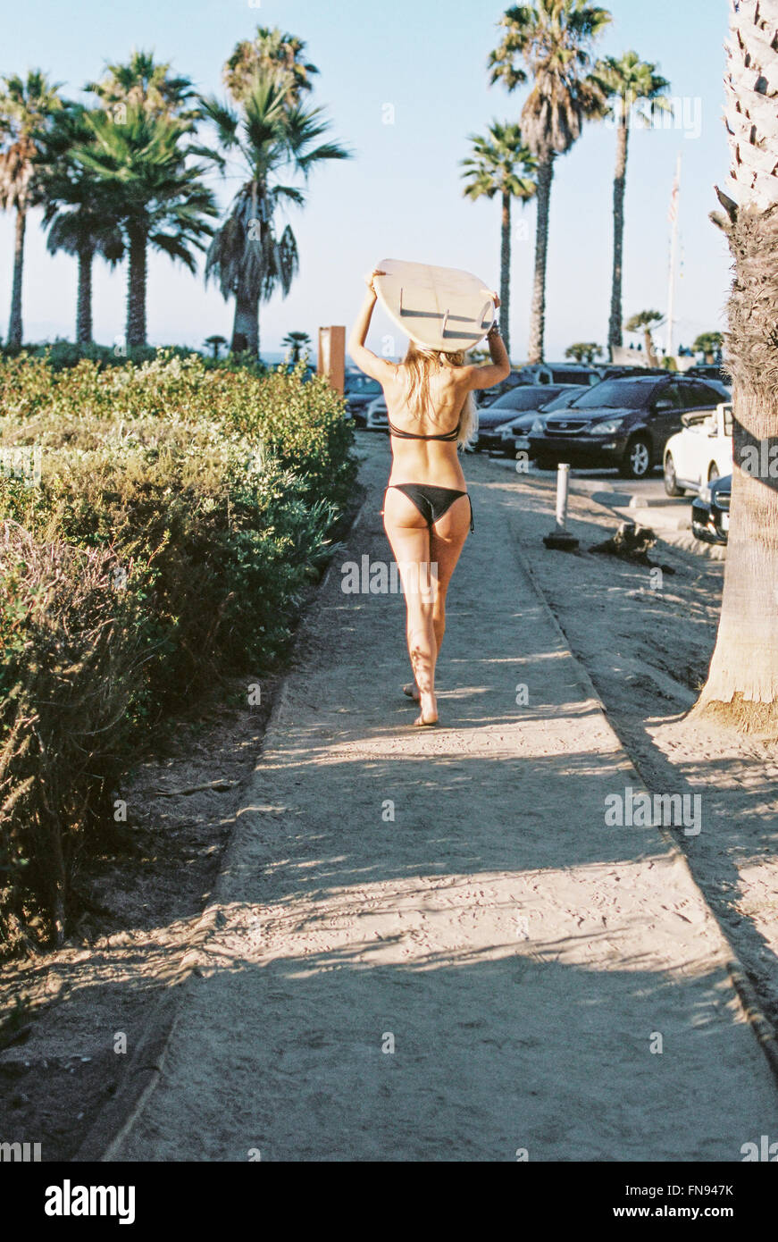 Una donna in bikini nero a piedi giù per un sentiero che porta una tavola da surf sulla sua testa. Foto Stock