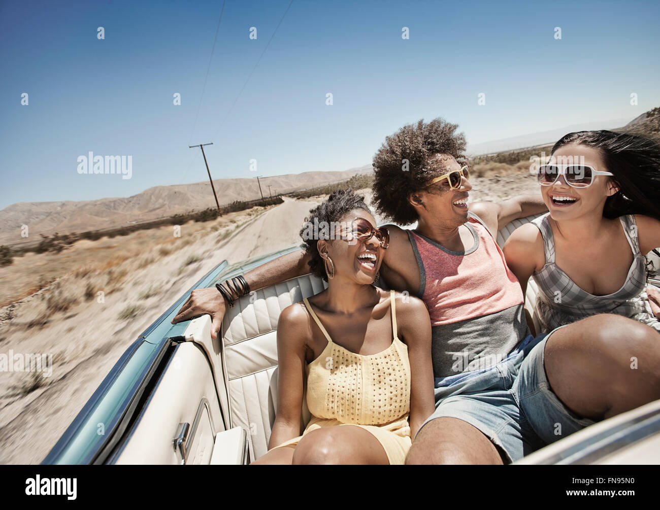 Tre giovani in un colore blu pallido autovettura convertibile, guida su strada aperta attraverso una piatta pianura a secco, Foto Stock