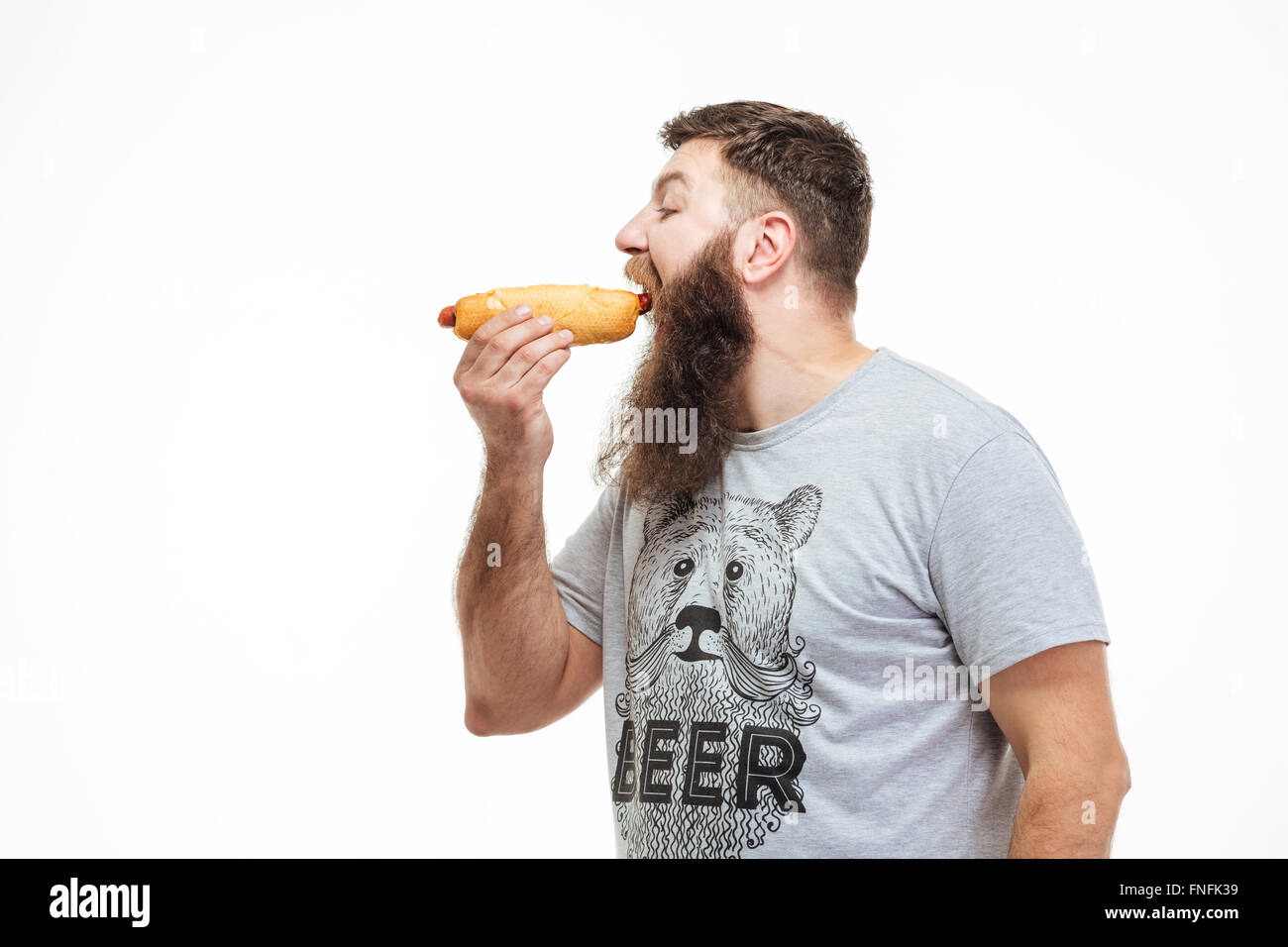 Profilo di uomo bello con la barba in piedi e mangiare hot dog su sfondo bianco Foto Stock