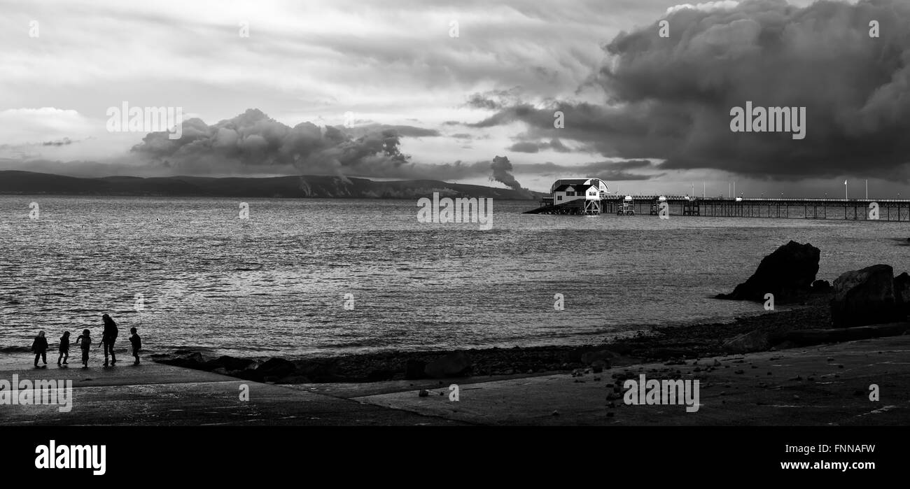 Monocromatico immagine in bianco e nero di famiglia giocando al calar del sole al tramonto sul foreshore con mumbles pier Galles, UK in background Foto Stock