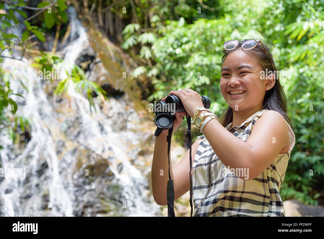 Bellissima fanciulla escursionismo è utilizzando un binocolo per guardare gli uccelli sorridente alla telecamera nella foresta tropicale vicino alle cascate Foto Stock