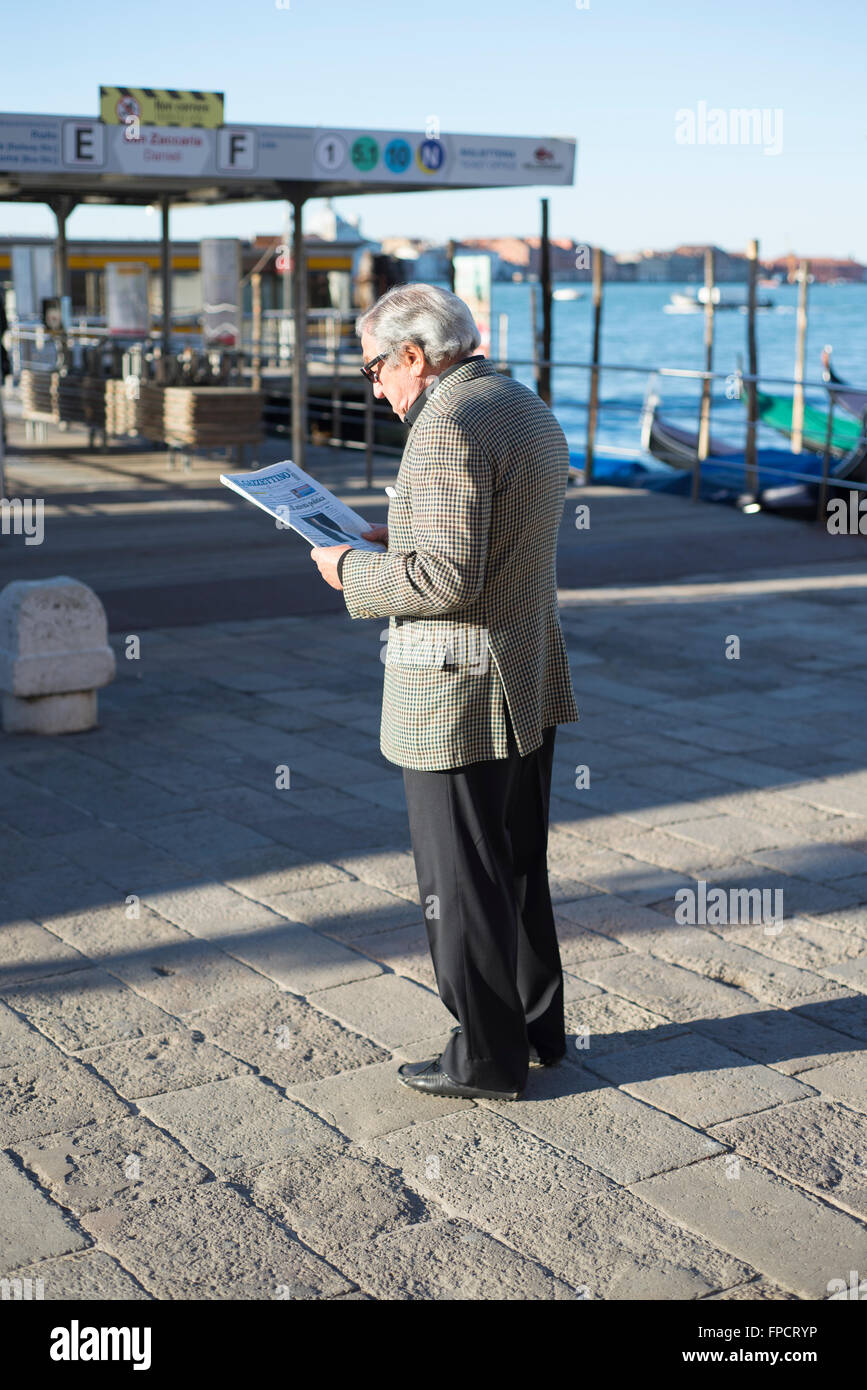 Uomo anziano leggendo il giornale di fronte al molo del traghetto sulla Riva degli Schiavoni quayside a Venezia in una giornata di primavera Foto Stock
