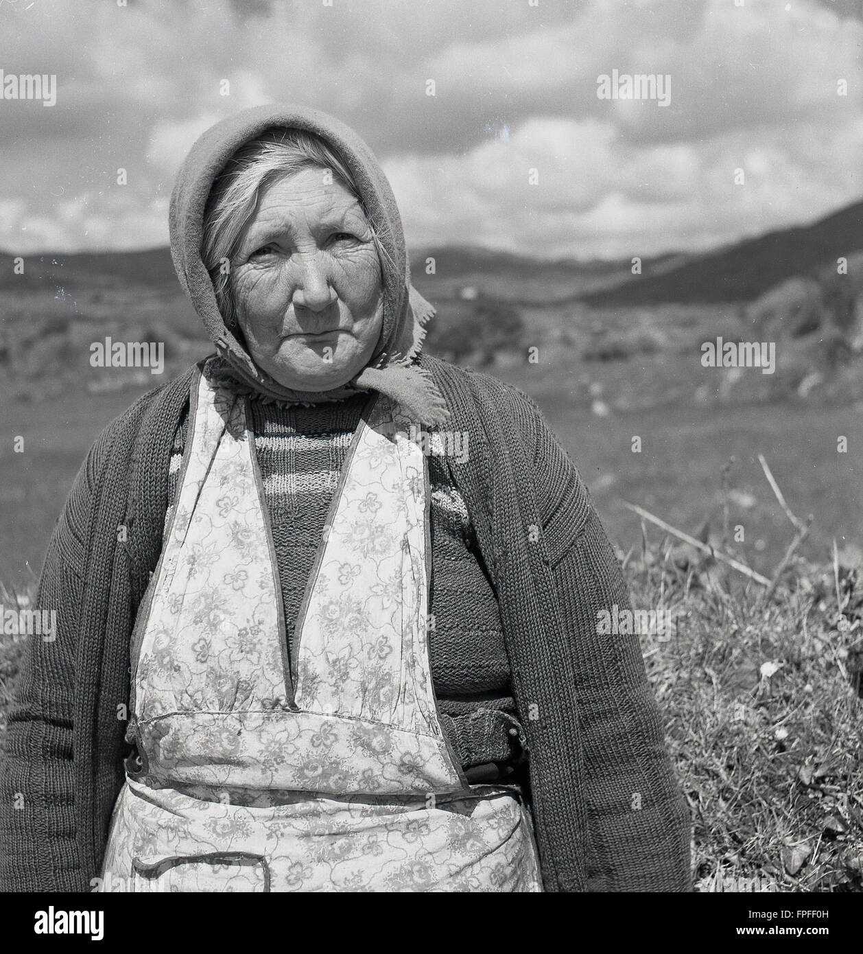 Anni '50, storico, un ritratto di J Allan Cash di una donna di campagna irlandese matura in una pinafore, cardigan e un velo, seduto da un hedgerow in campagna, nell'Irlanda occidentale. Foto Stock
