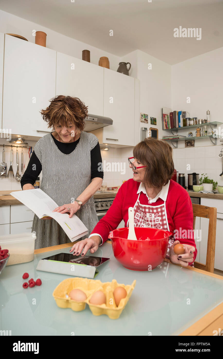 Due senior donna confrontando ricetta su tavoletta digitale e libro di cucina , Monaco di Baviera, Germania Foto Stock