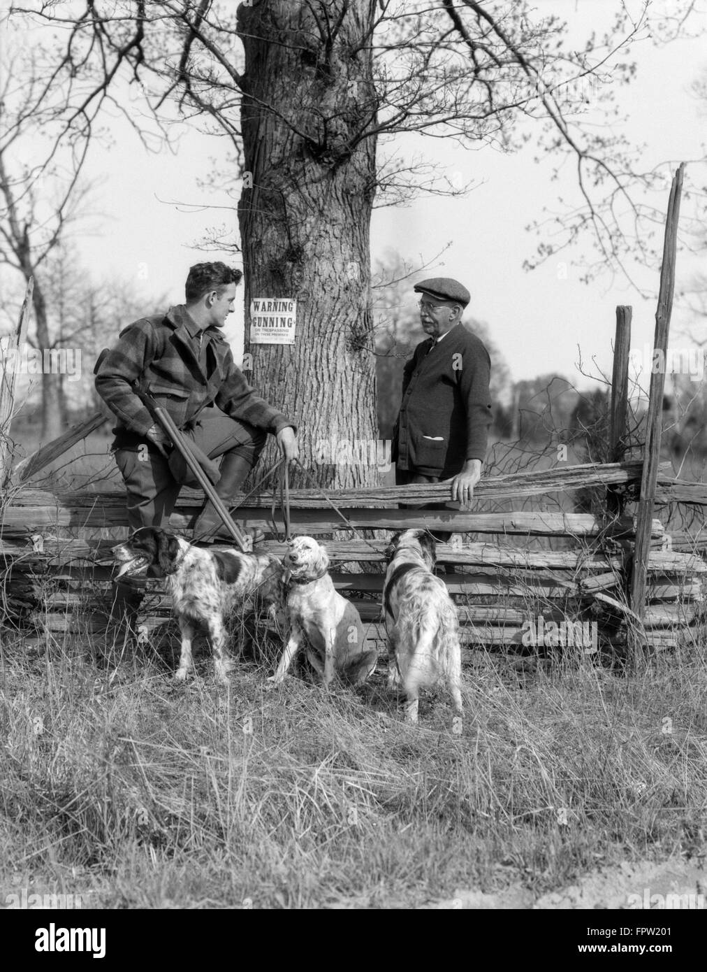 1920s senior uomo che parla all uomo giovane cacciatore con colpo di pistola e tre cani da caccia POSTER pubblicato su albero recinto di trasgressione Foto Stock