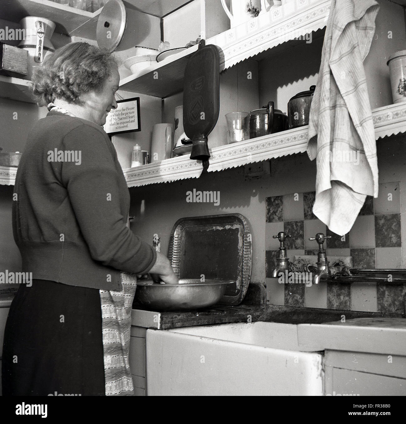 Anni Cinquanta storiche, una donna che indossa un cardigan sopra un grembiule in piedi con le mani in un recipiente stagno preparare cibi, accanto a un dissipatore di maggiordomo nella piccola cucina. Foto Stock