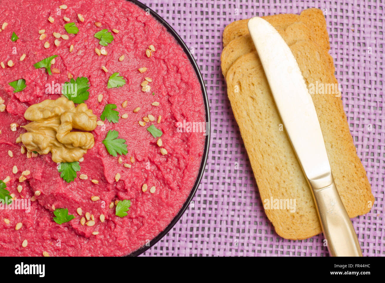 Hummus di barbabietole in una ciotola nero, un coltello d'oro e tre fette di pane su sfondo viola Foto Stock