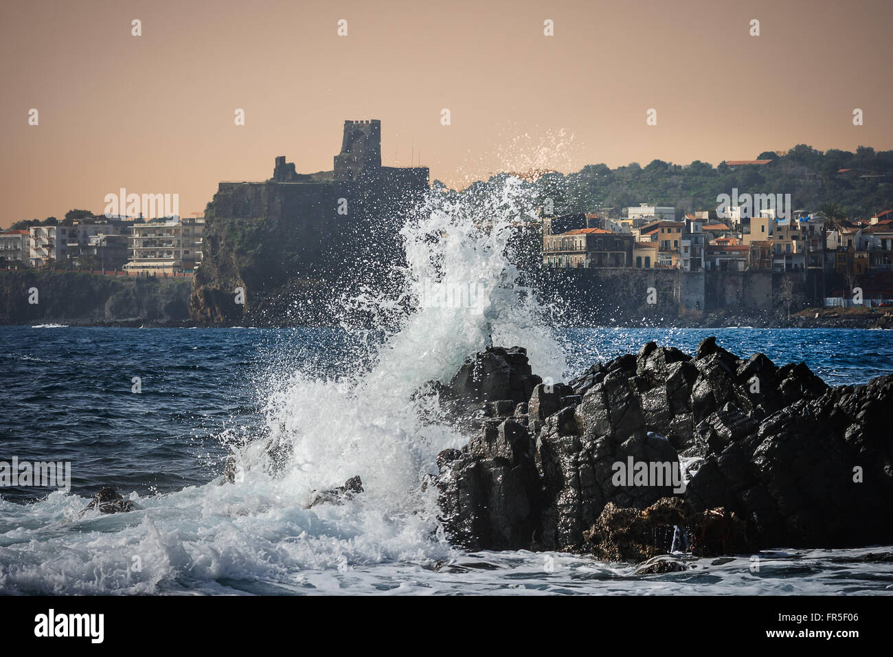 Seascape con una forte ondata contro una roccia e il castello normanno di Acicastello in background, Catania, Sicilia Foto Stock