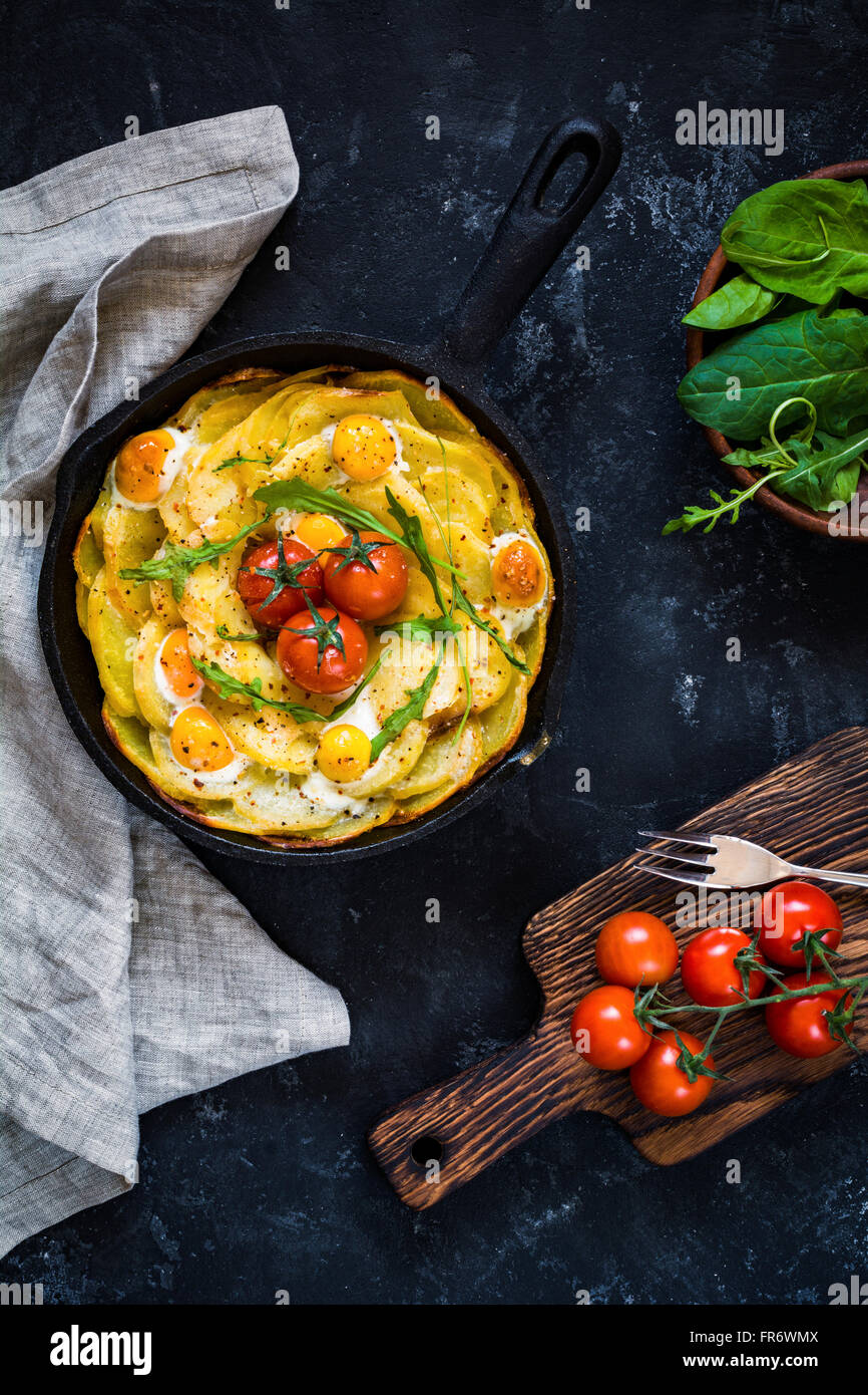 Colazione frittata di patate con spinaci, pomodorini e formaggio in padella. Tabella vista dall'alto su sfondo nero. Still Life Foto Stock
