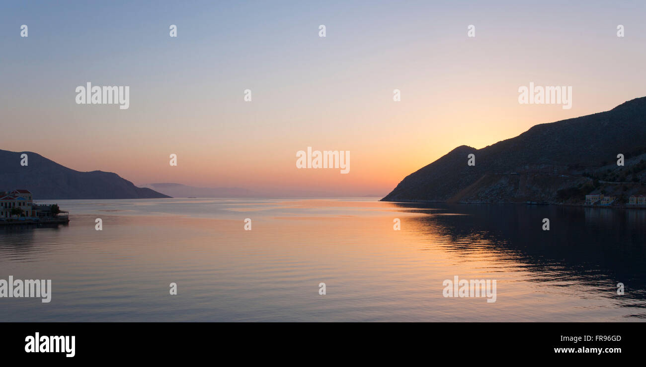 Gialos, Symi, Egeo Meridionale, Grecia. Vista panoramica attraverso le tranquille acque della baia di Harani, alba. Foto Stock