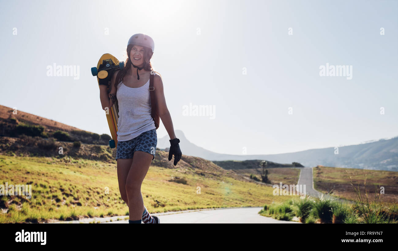 Felice giovane donna a piedi con un longboard sulla strada di campagna. Skater professionista all'aperto in una giornata di sole. Foto Stock