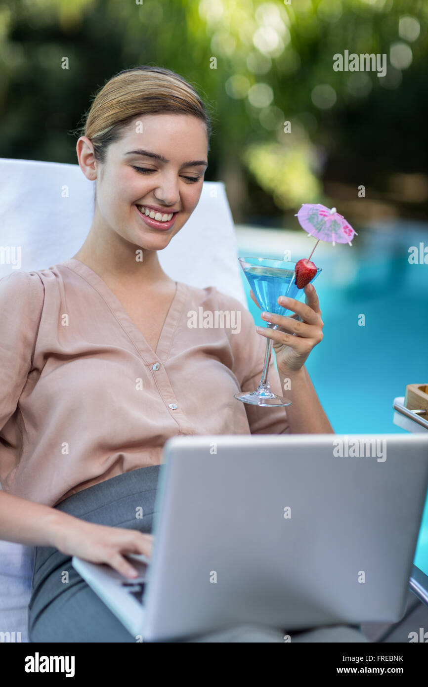 Imprenditrice tenendo un bicchiere di martini durante l'utilizzo di laptop Foto Stock