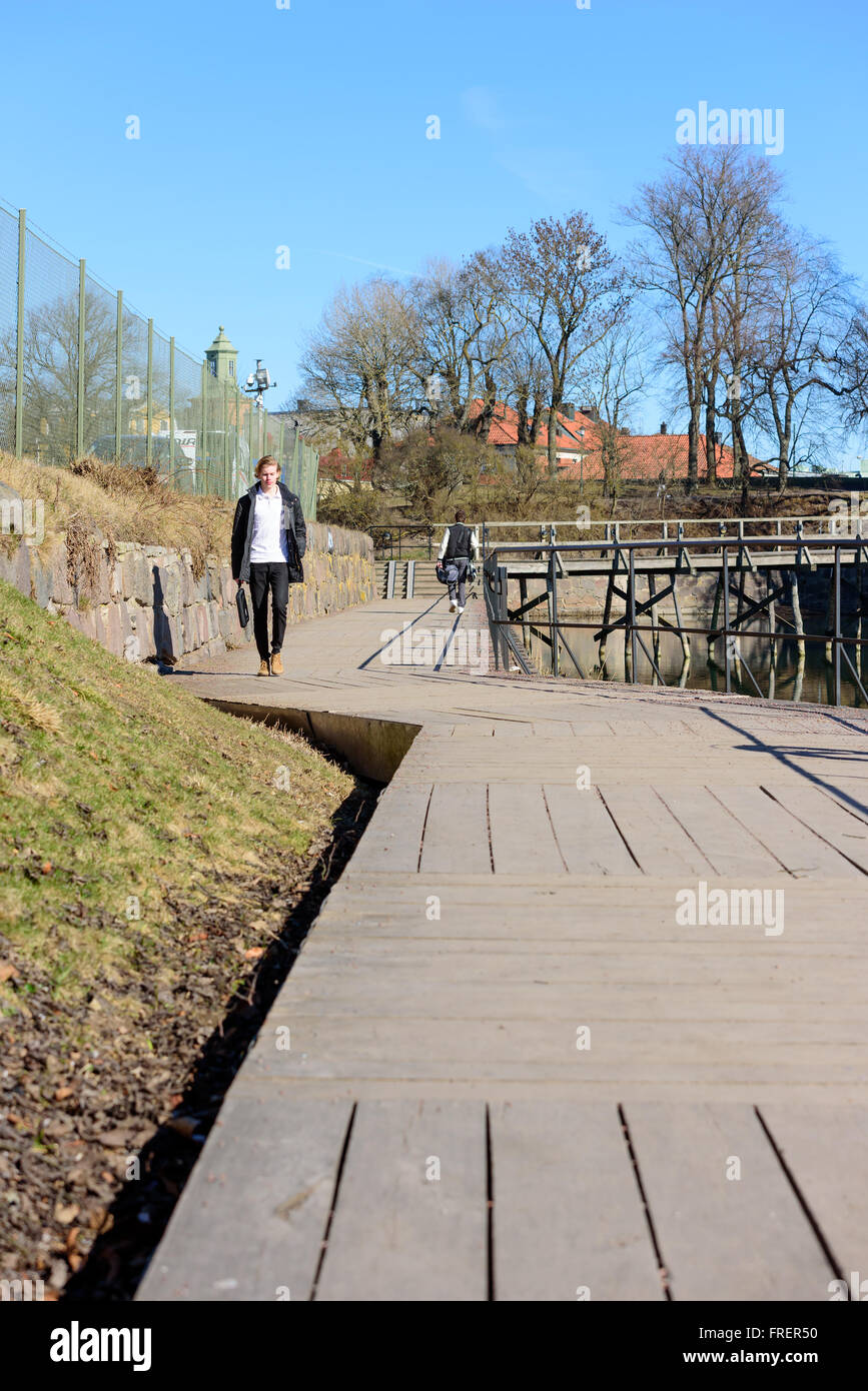 Kalmar, Svezia - 17 Marzo 2016: un giovane adulto a piedi verso di voi su una passerella di legno al di sotto di un recinto reticolare. Meteo è soleggiato Foto Stock