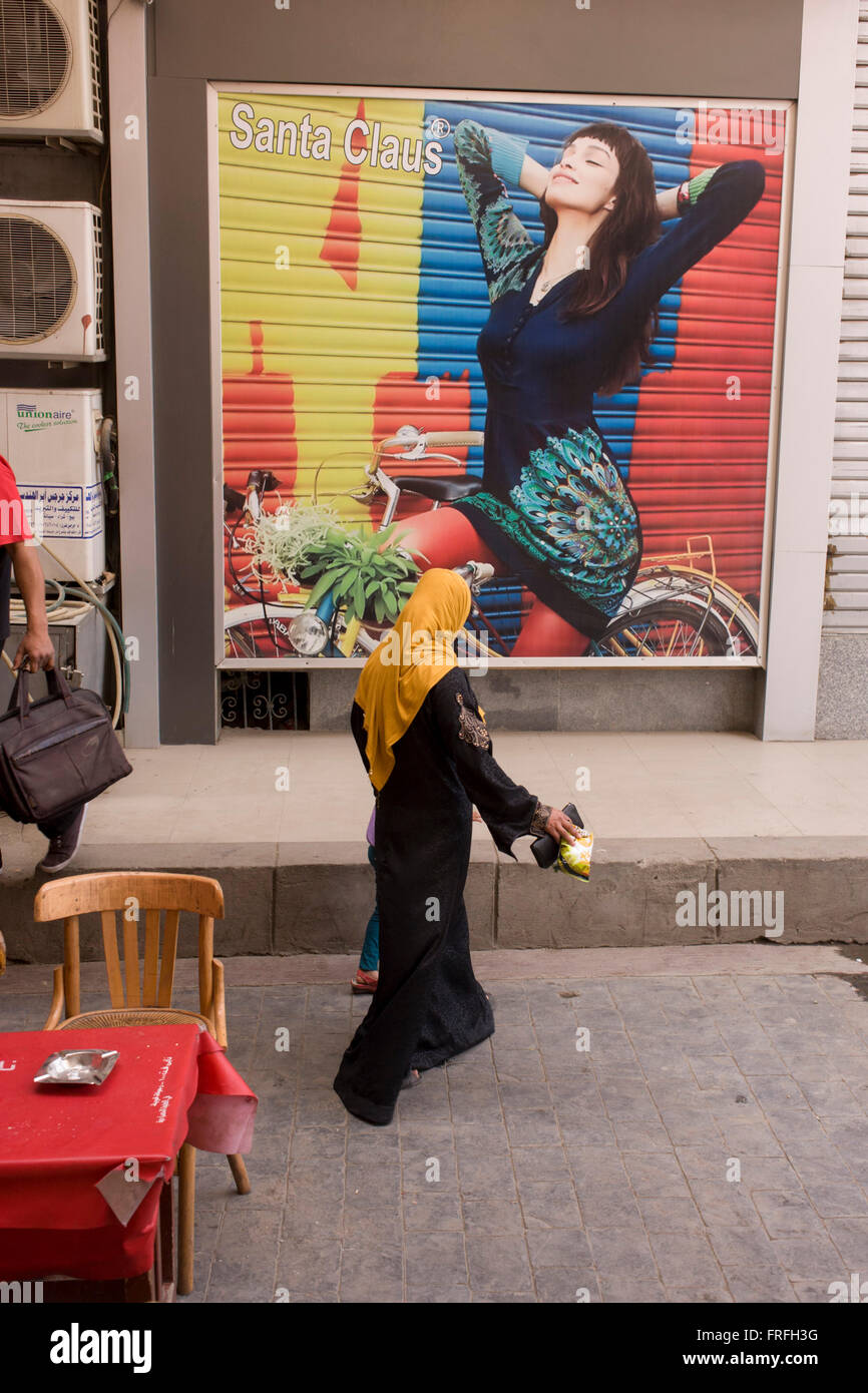 Tradizionalmente un vestito donna cammina sotto un poster per eleganti abiti arabi nella parte moderna della città di Luxor, la Valle del Nilo in Egitto. Foto Stock