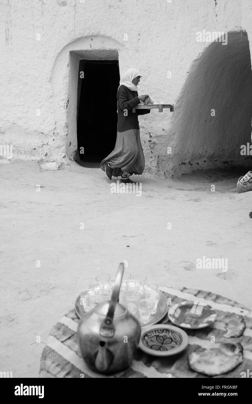 Donna berbera in uscita di una grotta tenendo un vassoio con il cibo mentre al primo piano è evidente una tabella con una teiera e g Foto Stock