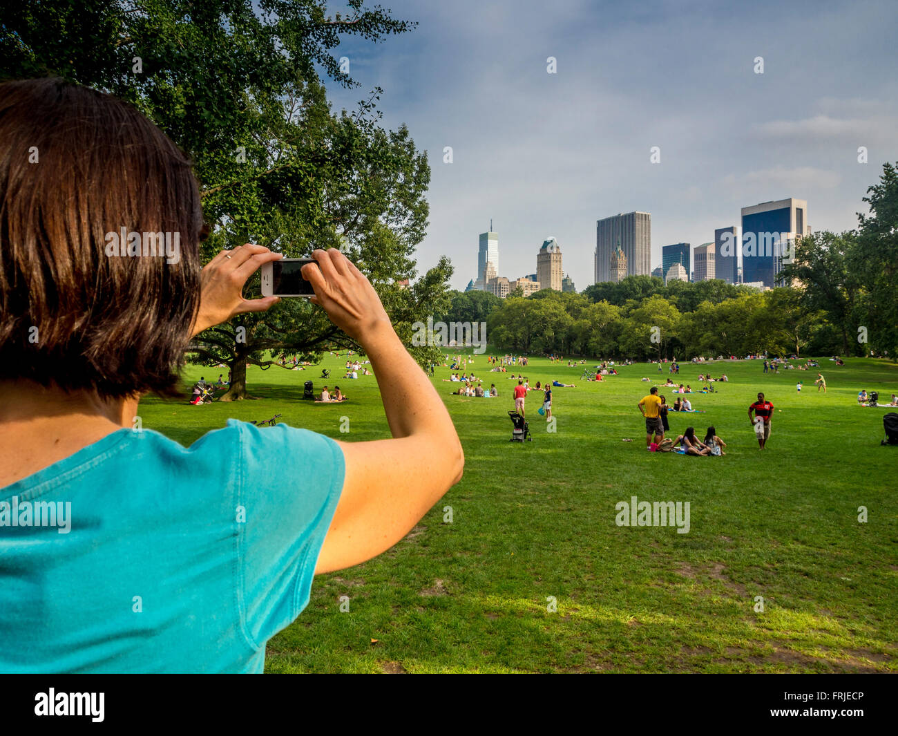 Donna che utilizza uno smartphone per scattare una foto di persone che si rilassano sull'erba di Central Park, New York City, con lo skyline di Manhattan sullo sfondo. Foto Stock