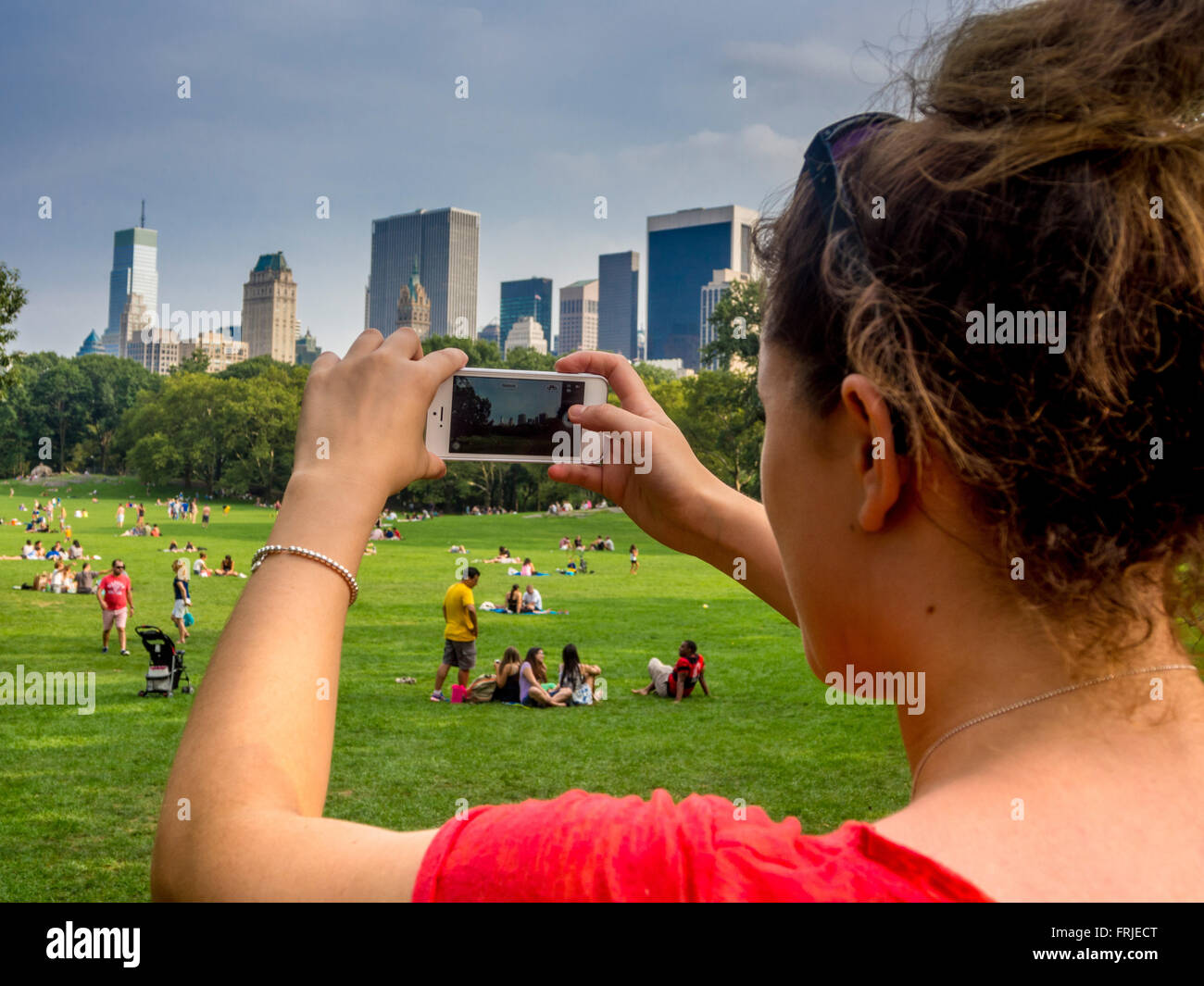 Donna che utilizza uno smartphone per scattare una foto di persone che si rilassano sull'erba di Central Park, New York City, con lo skyline di Manhattan sullo sfondo. Foto Stock