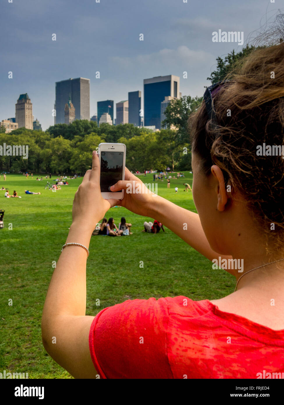Donna che utilizza uno smartphone per scattare una foto di persone che si rilassano sull'erba di Central Park, New York City, con lo skyline di Manhattan sullo sfondo. Foto Stock