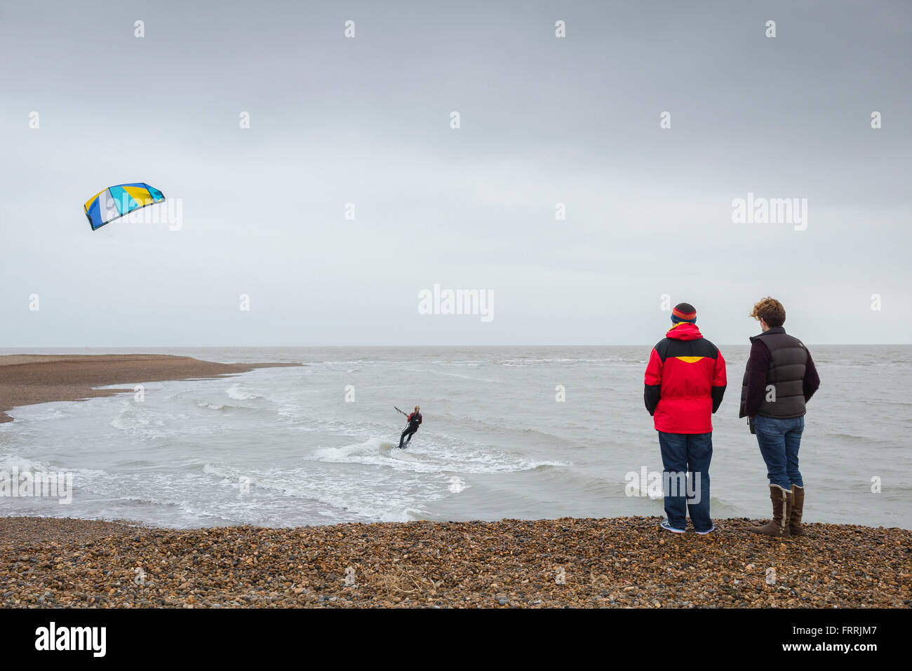 La gente guarda il kite surf, vista posteriore di una coppia in piedi su Hollesley Beach sulla costa di Suffolk guardando un kite surfer pattinare la costa, Inghilterra Foto Stock