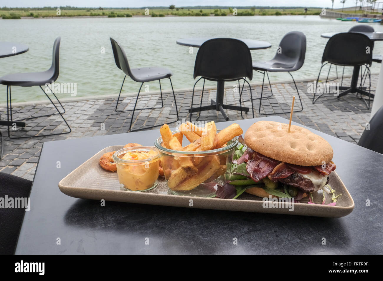 Hamburger di manzo su una piastra quadrata su una tavola nera, con patatine fritte, dip e anelli di cipolla, servita in un caffè all'aperto dall'acqua Foto Stock