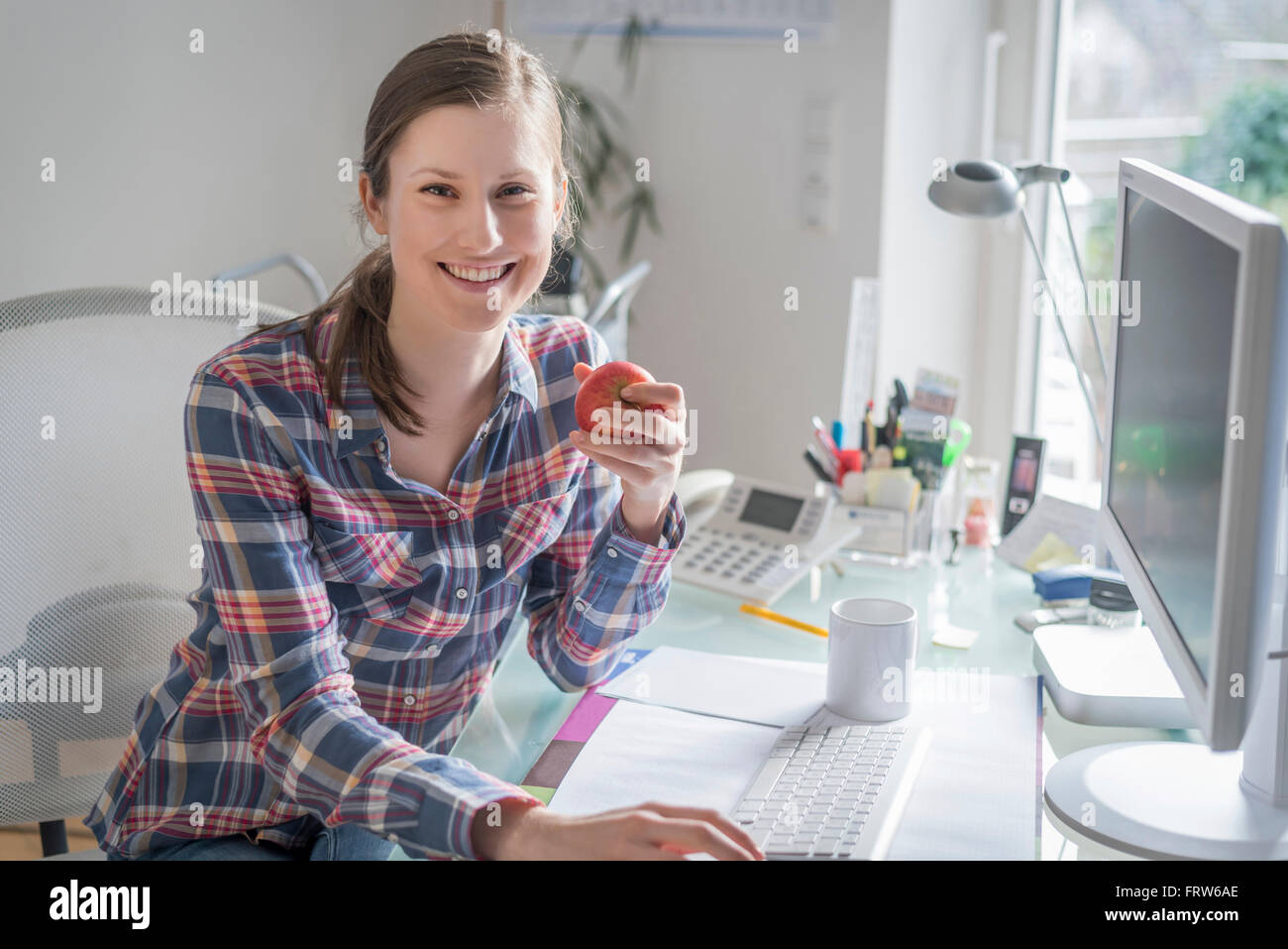 Ritratto di sorridente giovane donna alla scrivania in ufficio tenendo un Apple Foto Stock