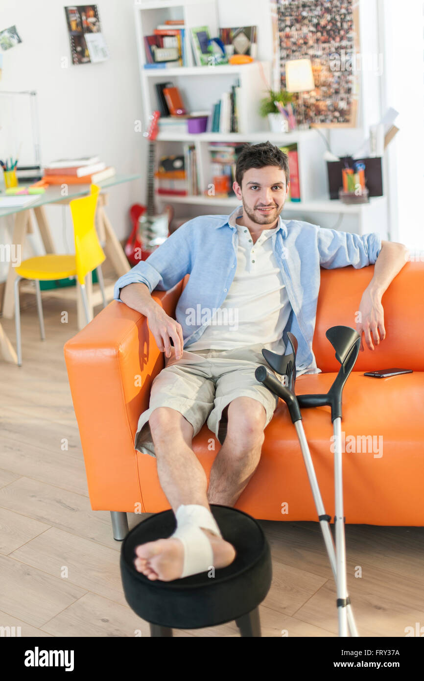 Giovane uomo con piede bendato seduto su un divano Foto Stock