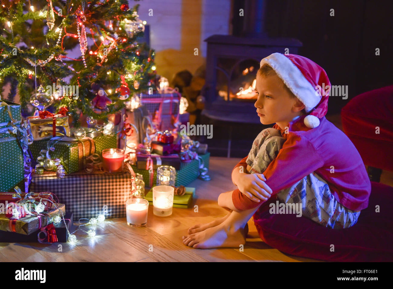 La Notte Di Natale.La Notte Di Natale Un Ragazzino Con Un Cappello Da Babbo Natale Seduto Su Un Cuscino Vicino Alla Stufa A Legna Nella Parte Anteriore Di Un Albero Di Natale Illuminato Foto Stock