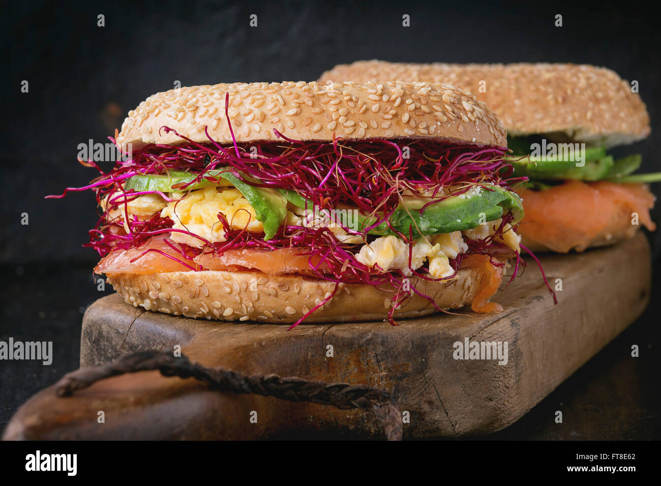 Due bagel con salmone salato, spinaci, germogli di barbabietola, avocado e uova strapazzate sul tagliere di legno su un vecchio legno nero Foto Stock