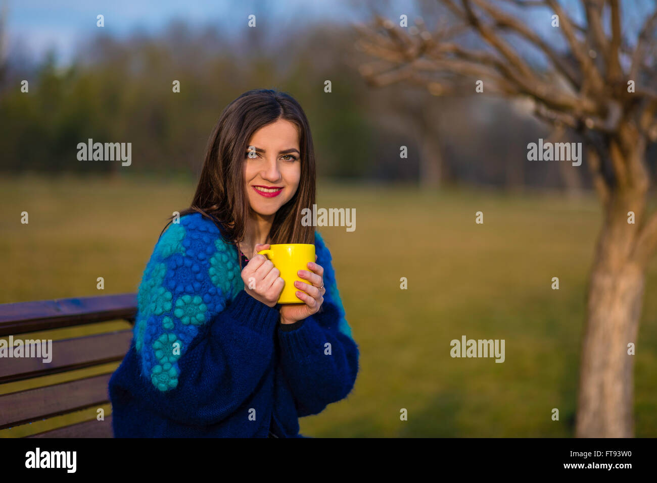 Una ragazza adolescente è seduta su una panchina in un parco e tenendo in mano un bicchiere di colore giallo con due mani. Foto Stock