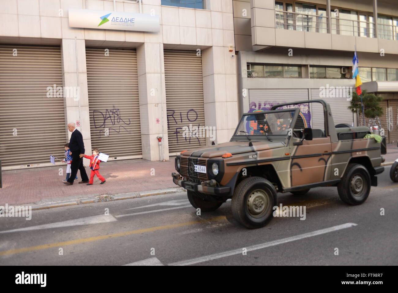 Atene, Grecia. 25 Mar, 2016. Un uomo con i suoi nipoti passando da Syggrou Avenue, in cui i veicoli militari che aspettano l'inizio della sfilata, sono parcheggiate da parte del personale. Indipendenza greca il giorno che è una festa nazionale si celebra ogni anno in Grecia per commemorare l inizio della guerra di indipendenza greca nel 1821 contro l'Impero Ottomano. Nella capitale della Grecia ad Atene un enorme parata militare si svolge come omaggio per il giorno speciale. Credito: Dimitrios Karvountzis/Pacific Press/Alamy Live News Foto Stock