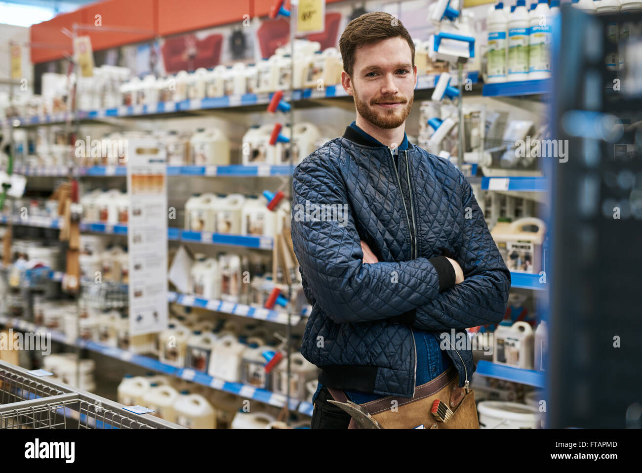 Fiducioso tuttofare in un negozio di ferramenta in piedi tra le rastrelliere di prodotti con bracci ripiegati sorridente verso la telecamera Foto Stock