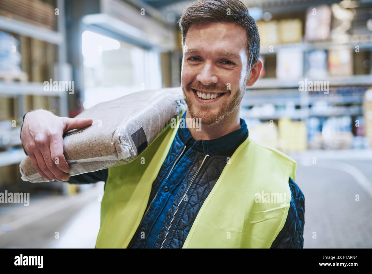 Gentile felice lavoratore del magazzino in un GIUBBETTO ALTA VISIBILITA in piedi nel magazzino con una sacca di prodotto sulla sua spalla smili Foto Stock