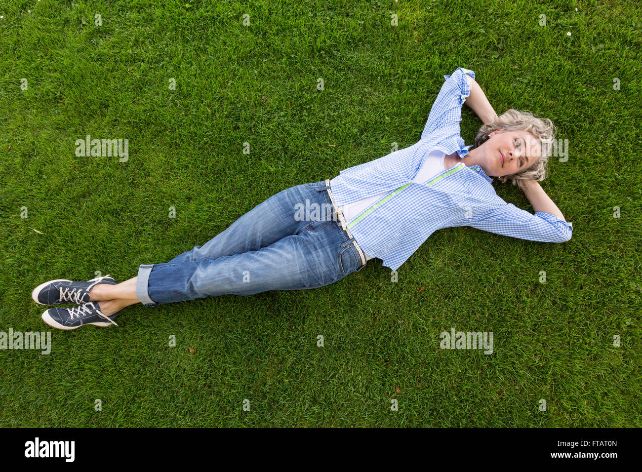 Donna di mezza età in casual abbigliamento weekend rilassante sull'erba in un cortile o parco. Lei è sorridente con una felice, felice espressione. Foto Stock