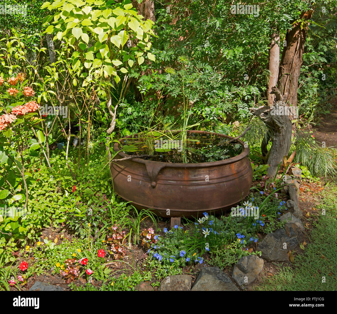 Spettacolare giardino con grande calderone arrugginito acqua caratteristica con Rosso, blu, bianco fiori & fitta verde smeraldo di arbusti dietro Foto Stock
