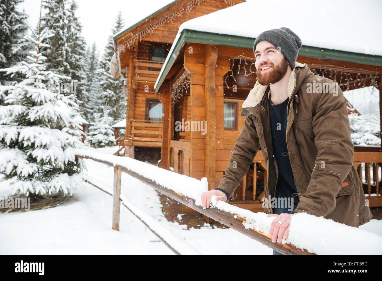 Ritratto di Allegro bello uomo barbuto in piedi nella parte anteriore del cottage di legno nella foresta di inverno Foto Stock