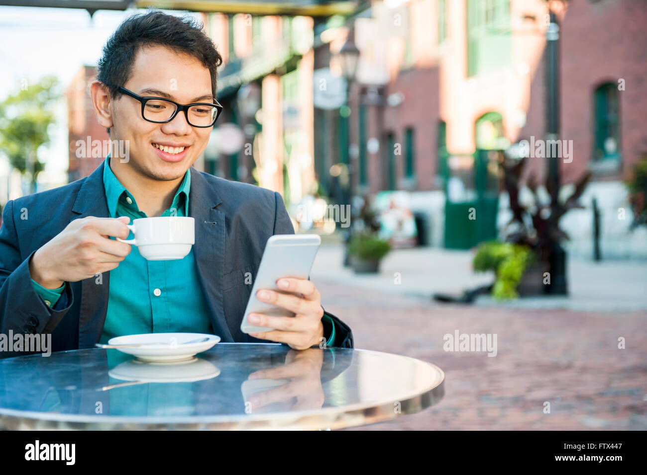 Giovane uomo asiatico in business abbigliamento casual seduto e sorridente in un rilassante outdoor cafe di bere caffè mentre utilizzando mobile Foto Stock
