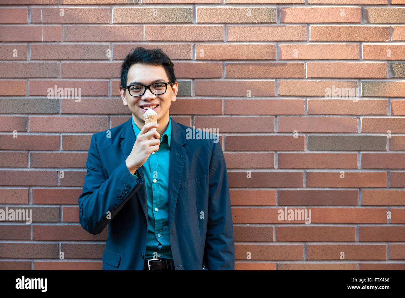 Sorridente giovane uomo asiatico nel suo ventenne godendo di gelato mentre sta in piedi vicino a un muro di mattoni al di fuori su una pausa Foto Stock