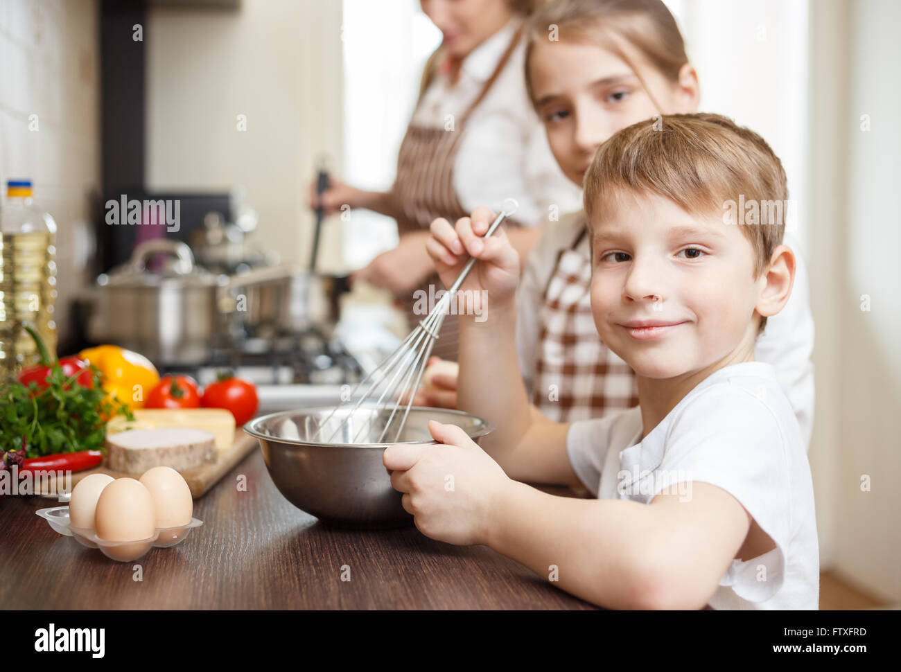 Sorridente piccolo ragazzo con mamma e mia sorella sbattere le uova nella ciotola sul tavolo. Cucina di famiglia dello sfondo. Foto Stock