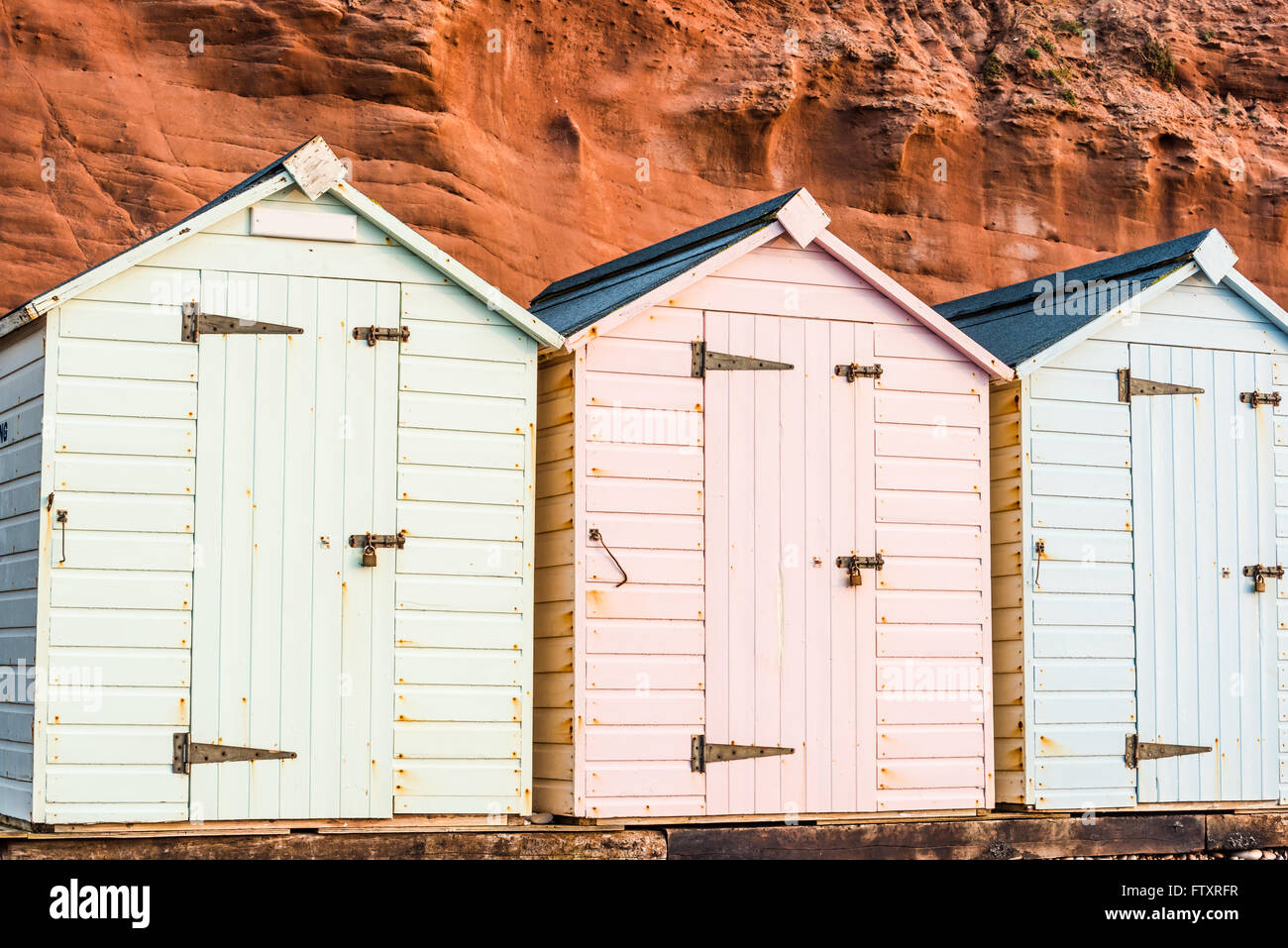 Beach Hut fila in colori pastello, roccia rossa sfondo, South Devon, Regno Unito Foto Stock