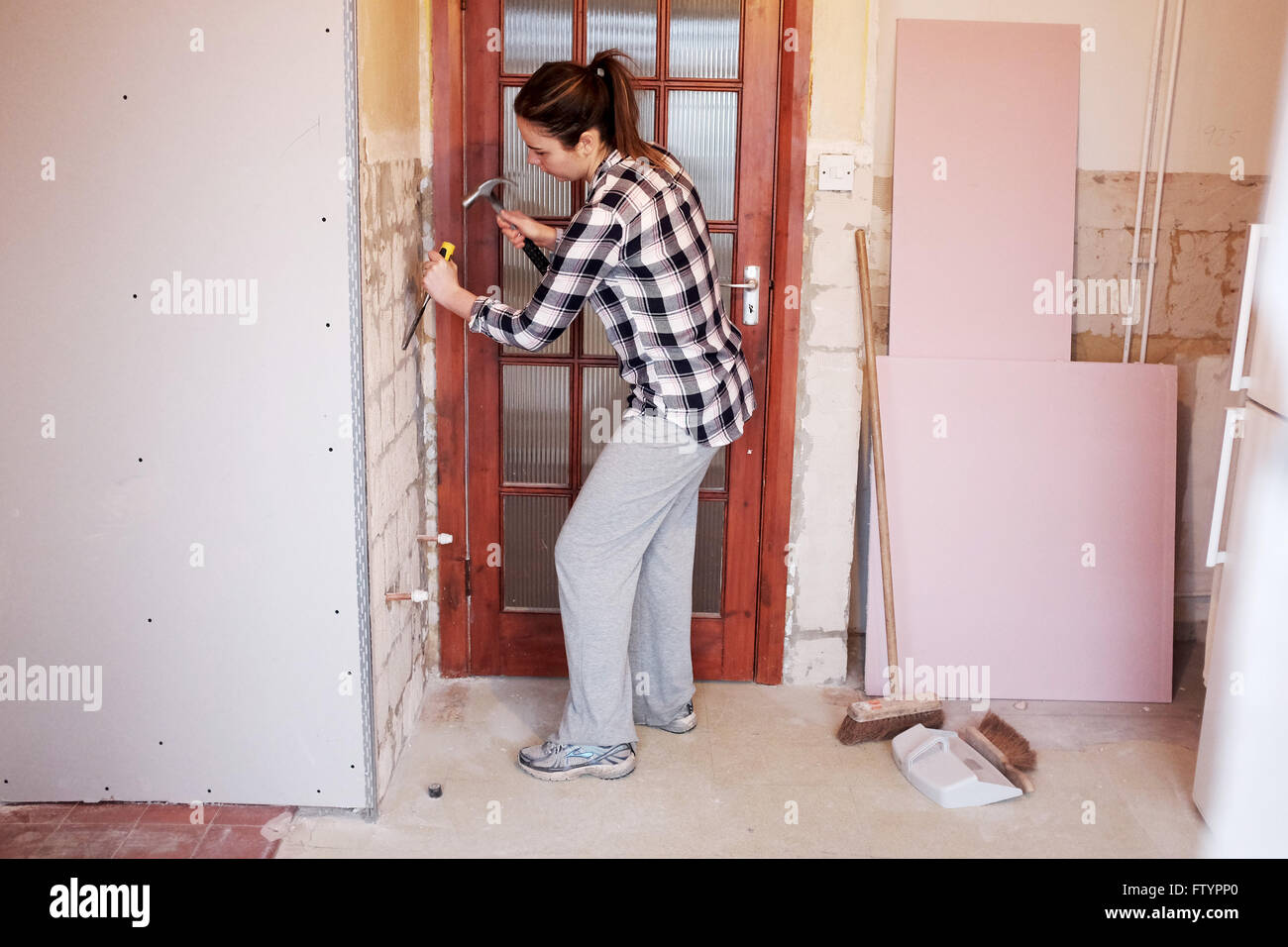 Giovane donna con un martello e uno scalpello di decollare piastrelle a muro in cucina nella sua 20s facendo DIY al suo bungalow home Foto Stock