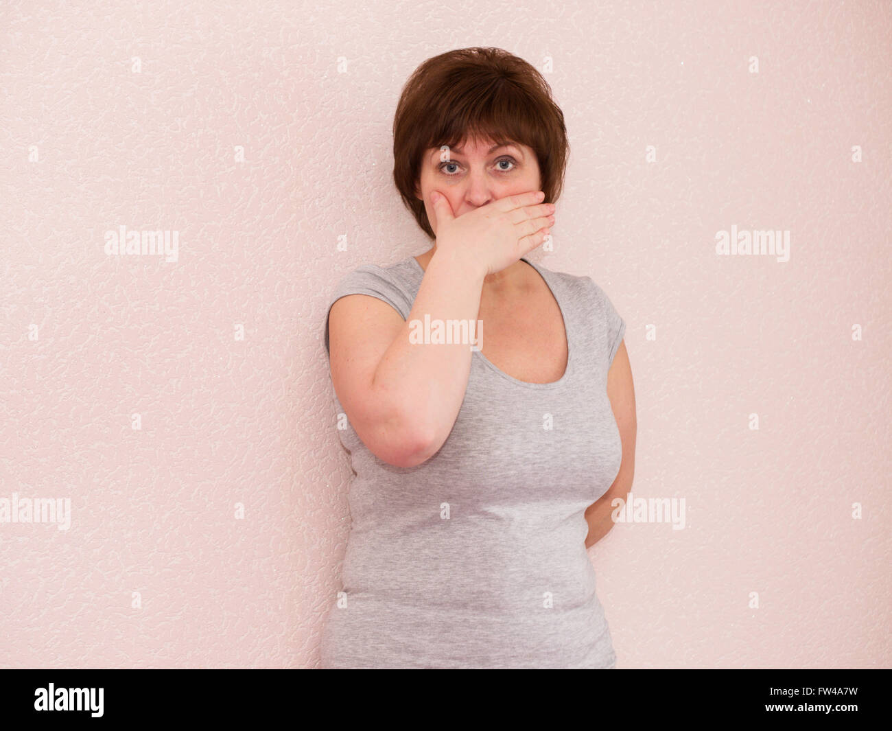 Ritratto attraente donna matura con scioccato, ansioso di espressioni facciali, che copre la bocca con la mano su sfondo semplice Foto Stock