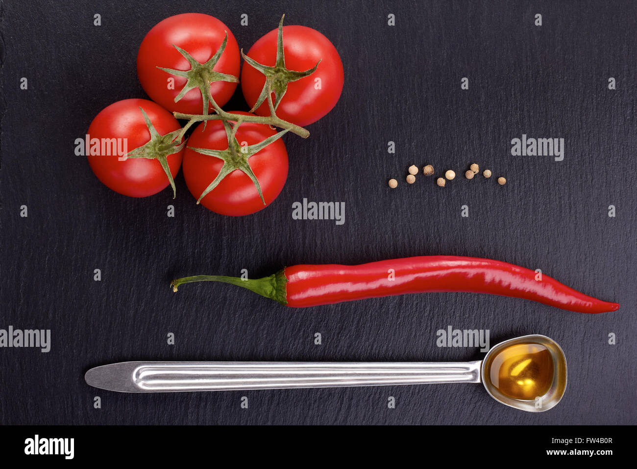 Grappolo di pomodoro, peperoncino, un cucchiaio di olio di oliva in pietra scura sullo sfondo. Foto Stock