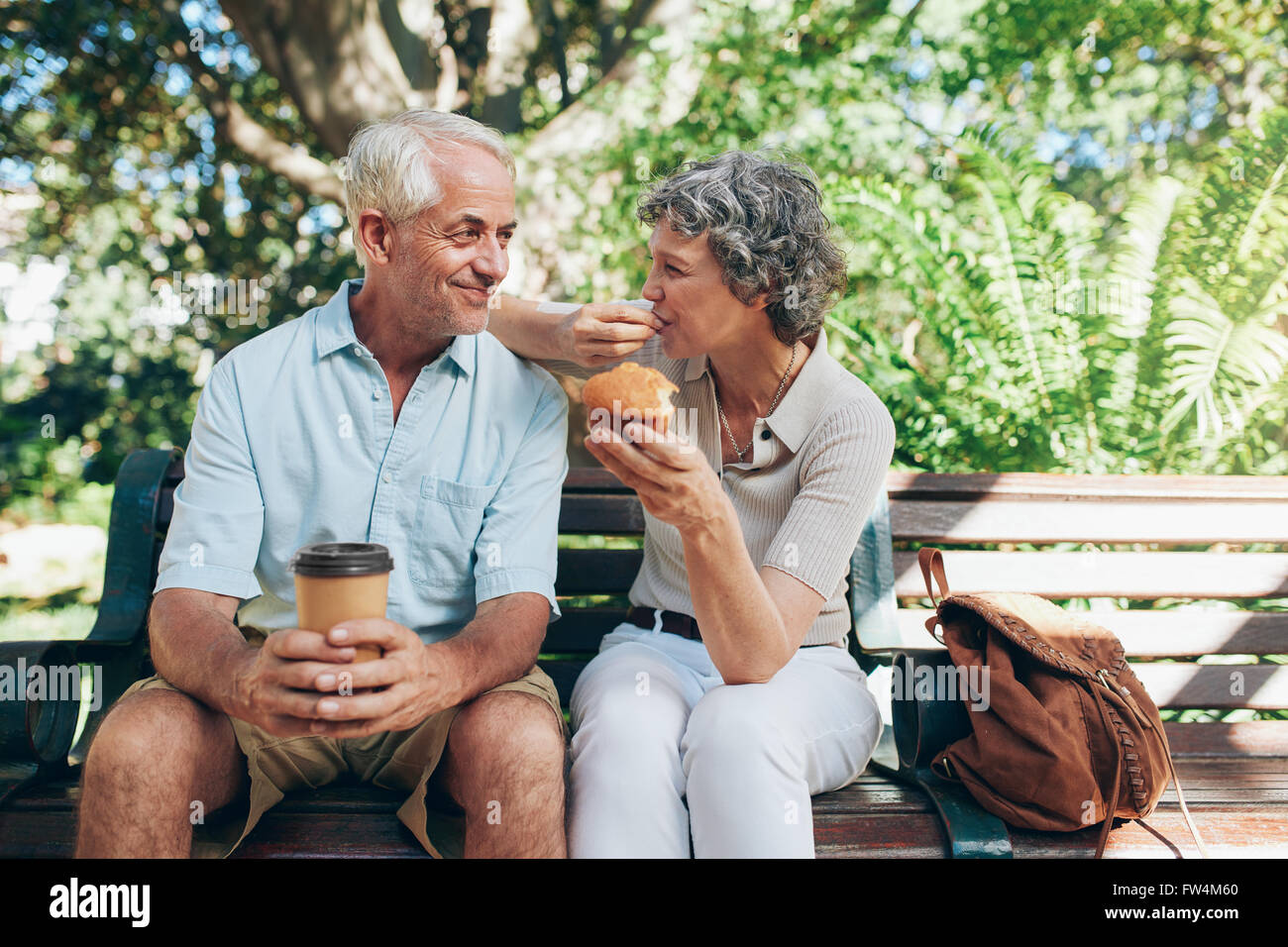 Amorevole coppia senior seduta su una panchina nel parco con caffè e muffin. Tourist relax all'aperto su una panchina nel parco. Foto Stock