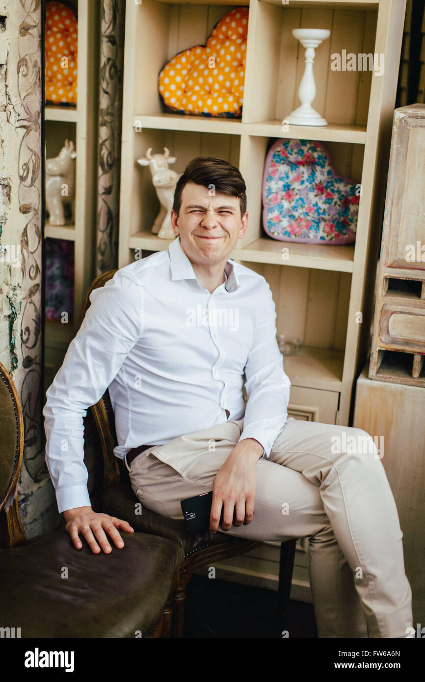 Ritratto emotivo di sorridere bello crazy hipster camicia bianca seduto su una sedia vintage con uno smartphone in stile retrò inter Foto Stock